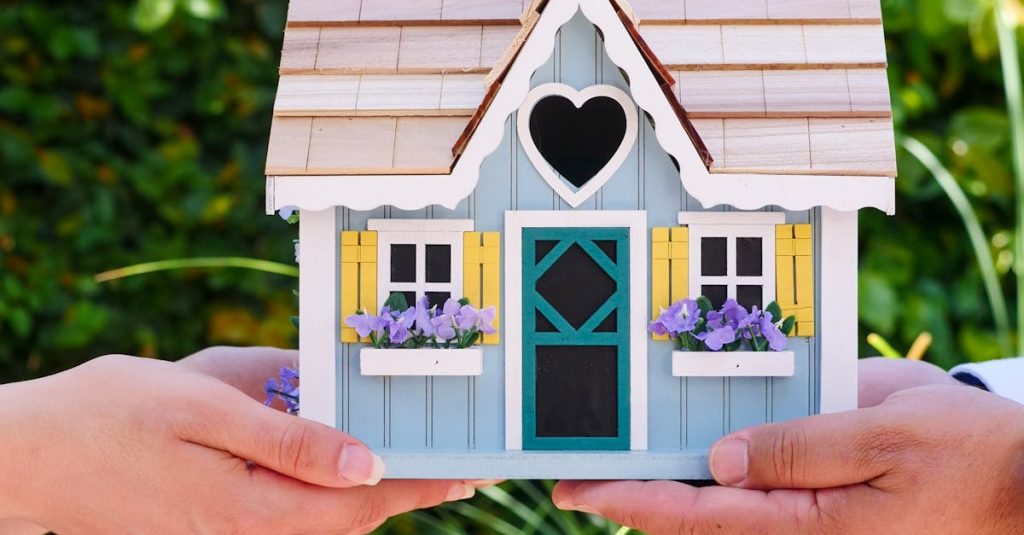 Two hands hold a colorful miniature house outdoors, symbolizing real estate and home ownership.